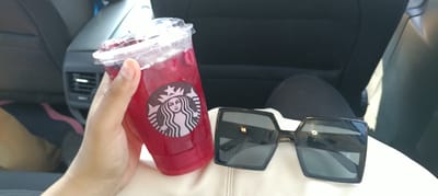 A hand holds a Starbucks iced beverage with a red liquid and the iconic siren logo. Next to the drink, a pair of oversized, dark-lensed sunglasses with a thick black frame rests on a white surface. The background shows the interior of a car, with a dark-colored car seat and a vent visible. The lighting suggests it is daytime. The image appears to be taken from the perspective of someone sitting in the car, likely during a drive. The overall mood is casual and relaxed, possibly indicating a personal outing or travel. The Starbucks logo is clearly visible on the cup. There is no other legible text in the image.