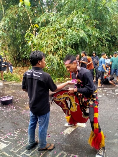 The image captures an outdoor scene on a wet, dark grey paved surface in Kedawung, Indonesia. In the foreground, a male on the left is viewed from the back and left profile, wearing a black short-sleeved shirt with white text "Seni Barongan Pusat Madiun" on the back, blue jeans, and black flip-flops. He has a lit cigarette in his mouth and extends his left hand forward. To his right, another male, dressed in traditional performance attire, faces left in mid-action. This individual wears a dark long-sleeved top, a multi-patterned sarong-like garment (red, yellow, brown, black), black leggings, and red and yellow faux fur adornments on his lower legs. He has a red and black patterned headband and holds a large, red, yellow, and black painted horse-head prop (kuda lumping) in his right hand, while his left hand is raised toward the first male's extended hand.
The paved ground is covered with scattered pink flower petals. A black bucket containing more pink petals is positioned on the far left of the frame. Faint white chalk or paint outlines are visible on the pavement in the mid-ground. Several other individuals are present in the mid-ground and background, observing the scene. One person is visible on their hands and knees in the mid-ground right, wearing attire consistent with the performer. The background is dominated by a dense wall of green bamboo foliage. The lighting appears overcast.