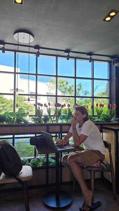 A Caucasian man is sitting at a small table by a large window, working on a laptop. He is wearing a white t-shirt and tan shorts, with black flip-flops. The window overlooks a lush green outdoor area with trees and flowers. The man has light brown hair and is wearing wireless earbuds. On the table in front of him is the laptop, a black case, and a cup of coffee. To his left, a dark green backpack is resting on a chair. The interior of the room appears to be a cafe or restaurant, with modern lighting fixtures on the ceiling and dark-colored furniture. The overall lighting suggests it is daytime, with bright sunlight streaming through the window. This scene is located in Choeng Mon, Thailand.