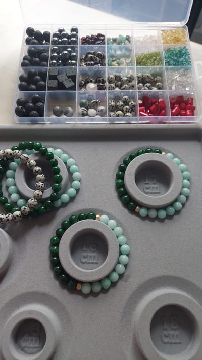 The image displays a top-down view of a crafting station, likely for jewelry making. In the upper portion, a clear plastic organizer with multiple compartments is filled with various types of beads, including black lava beads, metallic silver beads, dark red beads, green stones, and translucent white and yellow stones. Below the organizer, on a gray felt-like surface, are several beaded bracelets.
One bracelet, in the foreground left, is a mix of green and light blue beads with black and white speckled beads. Another bracelet, to its right and slightly behind, consists of green and light blue beads, with a single gold bead accent. Further to the right, a bracelet is composed entirely of light blue beads. There is also a bracelet visible in the upper left, made of green and black and white speckled beads. The gray surface has circular indentations with embossed measurements, such as "16 cm" and "18 cm," suggesting it is a display or measuring mat for bracelets.
The environment appears to be indoors, with soft lighting. There are no people visible in the image. The scene is focused on the materials and finished products of a jewelry-making activity. The location of Komsomolsk-on-Amur, Russia, is not discernible from the visual information provided in the image.