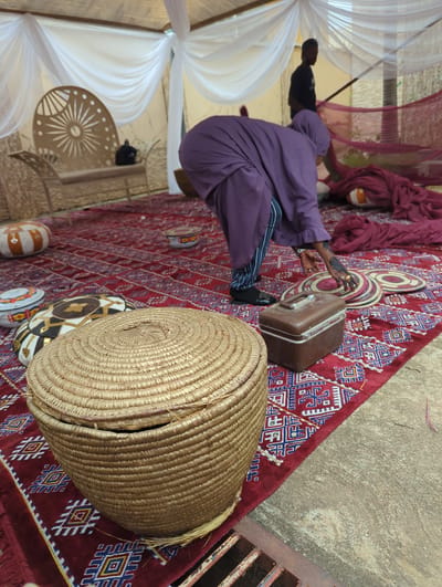 The image captures a vibrant cultural setting, likely an outdoor or semi-outdoor event in Unguwar Rimi, Nigeria. The scene is dominated by a rich, red patterned rug covering much of the ground.
In the foreground, a person dressed in a long, flowing purple garment with striped trousers is seated on the rug, facing away from the camera. Their left hand, adorned with intricate henna designs and rings, is visible near some smaller, round, woven decorative mats placed on the rug. Next to them is a vintage-style brown hard-shell suitcase. To the right, a large, round, lidded basket made of woven straw occupies the immediate foreground.
The surrounding environment features decorative elements such as white and maroon/red sheer fabrics draped, suggesting a temporary canopy or tent setup. In the background, another person wearing a black t-shirt can be seen standing. A small, ornate metal table with a sunburst pattern is also visible. The area beyond the rug appears to be a concrete or dirt ground, with a metal grate visible on the far right edge.
The presence of henna, traditional woven items, and the patterned rug strongly indicates a cultural celebration or ceremony, possibly a wedding or engagement, which is common in Nigerian traditions. The image appears to be taken during daylight hours. No discernible text is visible.