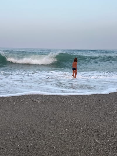A solitary child, seen from behind, stands in shallow surf on a dark pebble beach. The child, wearing dark swim trunks, faces a breaking wave. The water is a deep turquoise with white foam washing ashore. The sky is a pale, hazy blue, indicative of early morning or late evening. The background shows a serene, vast ocean with a large wave cresting. The foreground is dominated by the dark, coarse texture of the pebble beach. The overall atmosphere is tranquil and contemplative. The scene is located on the coast of Fuengirola, Spain.