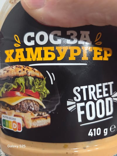 A person's fingers are visible at the top, holding a cylindrical food product container with a black label, captured in Stroevo, Bulgaria. The label features prominent Cyrillic text "СОС ЗА ХАМБУРГЕР" (Sauce for Hamburger) in white and orange. An illustration of a prepared hamburger with lettuce, tomato, cheese, and a patty is centered on the label. To the right, the English text "STREET FOOD" is displayed. A Nutri-Score indicator points to "D" on a green-to-red scale. The net weight "410 g e" is printed at the bottom right. The container's visible portion is a light, creamy orange color. A "Galaxy S25" watermark is present in the lower-left corner.