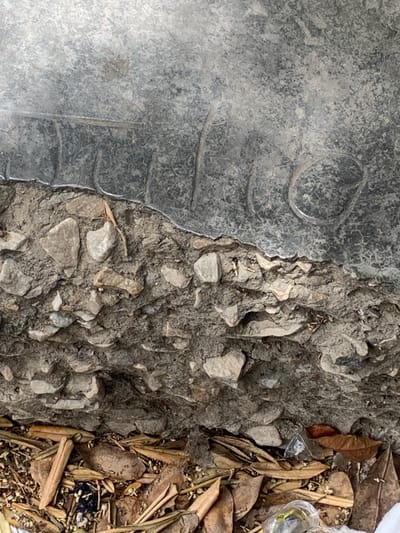 The image displays a close-up view of the ground's surface, likely outdoors given the presence of dry leaves and natural debris. The scene is divided into three distinct textures. On the left, a large, dark, and somewhat reflective surface dominates, exhibiting a smooth, almost metallic appearance with subtle circular and curved indentations. This surface appears to be a manufactured object, possibly a piece of plastic or metal.
Adjacent to this, in the center, is a rough, textured area of concrete or asphalt studded with various-sized grey stones. This section is uneven and appears weathered. Small, light-colored fragments are visible among the stones, some resembling bone fragments.
To the right of the concrete, the ground is covered with a layer of dry, fallen leaves and twigs, along with some indeterminate small objects and a small clear plastic container holding what appears to be green material. The overall impression is one of a neglected outdoor space.
No people are visible in the image, and no specific activity is taking place. The lighting suggests it could be daytime, with no strong shadows indicating a particular time. The location cues, such as the dry leaves and debris, are consistent with an outdoor environment that might be subject to wind or seasonal shedding. There is no visible text in the image. The context provided for Tacarigua, Venezuela, suggests a tropical or subtropical climate where such natural debris would be common.