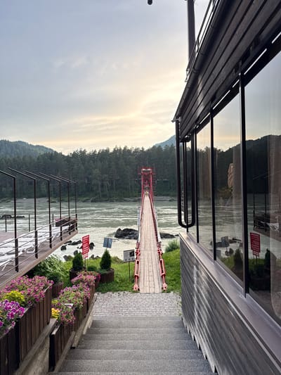 The image shows a view of a pedestrian suspension bridge over a wide river. The bridge is painted red and has a wooden walkway. On the right side of the image, a modern building with large glass windows is visible, reflecting the surrounding landscape. In the foreground, there are stairs leading down to a pathway lined with planters filled with purple and pink flowers. The pathway leads to a wooden deck with rows of white lounge chairs. The river is surrounded by a dense forest of pine trees, and mountains can be seen in the background. The sky is overcast with hints of sunlight breaking through. The location is identified as Barangol, Russia. There are no people clearly visible in the image. The scene appears to be a recreational area or resort.