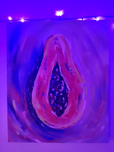 A square-format painting, depicting a lengthwise cross-section of a papaya fruit, is mounted on a light-colored wall in Mas, Indonesia. The artwork features vibrant pink and reddish-orange hues for the fruit's flesh, contrasting with a central cluster of dark, circular seeds. The background of the painting is composed of swirling brushstrokes in various shades of blue and purple. The entire scene is illuminated by a predominant blue-violet ambient light. Horizontally positioned above the painting, a black cord supports multiple small, glowing warm-white LED lights, which contribute additional illumination to the upper wall and ceiling area. No individuals, actions, or direct interactions are visible.