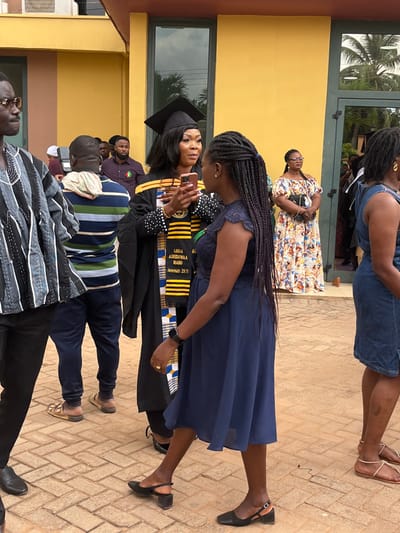 The image captures an outdoor scene in Madina, Ghana, on a paved area composed of red-brown interlocking pavers. A yellow-painted building with dark-framed glass doors and window panels, reflecting green foliage, forms the background.
In the foreground and mid-ground, multiple individuals are present. The central figure is a woman wearing a black academic gown and mortarboard cap, characteristic of a graduation ceremony. Her gown features white beaded cuffs, and she wears a black, yellow, and blue kente-patterned stole or sash with the text "LINDA ASEDUWAA MANU Jeremiah 29:11" visible. She holds a metallic pinkish-gold smartphone horizontally in front of her, pointing towards another woman.
This second woman, positioned to the right of the graduate, wears a dark blue dress with a lace-textured bodice and has long dark braided hair. She has a black wristband on her left wrist and a gold-colored ring on her left ring finger, and wears black low-heeled strap shoes. She appears to be interacting with the graduate.
To the far left, partially visible, is a man wearing a black and white striped shirt resembling Kente cloth and eyeglasses. In the mid-left background, a man facing away wears a blue, green, and white striped shirt and light-colored sandals. In the mid-background, near the building entrance, a woman wearing glasses and a long, multi-colored floral dress stands. Another woman, partially visible on the far right, wears a denim-like dress and light-colored sandals. Additional blurred figures are present throughout the background.