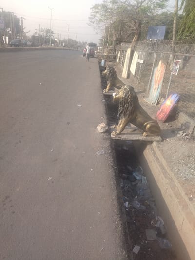 The image depicts a street scene in New Karu, Nigeria, located at coordinates 8.998 latitude, 7.571 longitude. There are three golden lion statues positioned on the edge of a drainage ditch, which is filled with debris. The statues are positioned along a paved road with a small sidewalk beside it. The road is empty of vehicles, though there are three people visible in the distance walking down the road. The image is taken from a low angle and appears to be looking down the road. There is a chain-link fence in the background, and several paintings or posters are hanging on the fence. The environment is dusty and dry. The scene suggests a combination of urban and rural settings.