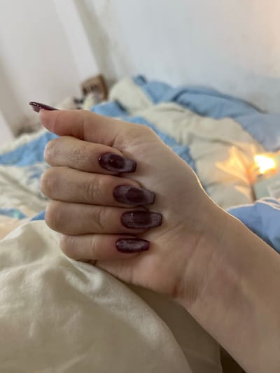 A hand with long, dark red nails is resting on a textured, light-colored surface, likely a blanket or bedding. The nails have a glittery or shimmering effect, possibly achieved with polish or a nail art technique. The hand is positioned in a way that displays the nails prominently. The background includes a blurred view of what appears to be a blue and white duvet cover. The lighting highlights the texture of the bedding and creates a soft glow on the hand.