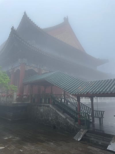 The image shows a traditional Chinese building shrouded in heavy fog. The structure features layered, tiled roofs, red pillars, and intricate decorative details. Adjacent to the building is a set of stairs and a stone wall. The scene is located in Yongding District, China.
