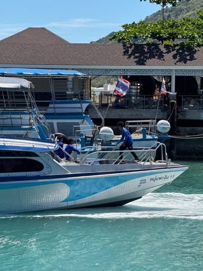 A medium shot captures a boat moving through turquoise waters under a clear blue sky. The boat, painted with blue and white stripes, has several people on deck. On the stern of the boat, two individuals in blue shirts are seated. Further forward, another person in a blue shirt and dark pants is seen walking. The boat bears the markings "6946 00165" and Thai script on its side. In the background, a building with a brown shingled roof and a white decorative facade is visible. Two Thai flags are displayed near the building. The surrounding environment appears to be a coastal area, likely a harbor or bay, given the presence of boats and the building which could be a pier or waterfront establishment. The water exhibits white foam trails from the boat's movement, indicating it is in motion. The overall scene suggests a sunny day in a tropical location. The specific location is Ban Haad Rin, Thailand.