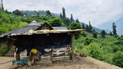 The image depicts a rustic dwelling in a hilly, verdant landscape, possibly in the vicinity of Shila, India. The structure features a dark, tiled roof with a conical section and an extended corrugated metal awning supported by wooden beams. The walls appear to be constructed from a combination of mud, stone, and possibly some salvaged materials, with blue and white tarps partially covering the front.

In front of the dwelling, a small garden area is visible with several potted plants and some wildflowers. Various tools and objects are scattered around, including a wheelbarrow, shovels, and a yellow bag. The ground is uneven and appears to be dirt or gravel.

The background is dominated by lush green hills covered in dense vegetation and trees, with a misty mountain range visible in the distance. The sky is overcast with scattered clouds. Several other scattered houses and structures are visible on the hillsides in the mid-ground, suggesting a rural settlement. The overall impression is one of a remote, natural setting.