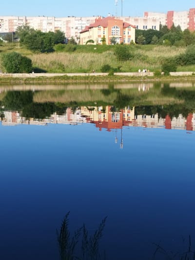 The image shows a calm body of water in Novyi Rozdil, Ukraine, reflecting the buildings and greenery along its edge. The buildings are a mix of multi-story apartment blocks and a smaller, reddish-orange building with a distinctive architectural style, possibly a public building or a unique residential structure. The reflection is quite clear, showing the colors and shapes of the structures and the vegetation. On the embankment, there are trees and tall grasses. The sky is clear and blue. In the foreground, the dark blue water has a smooth surface, with some reeds or tall grass visible at the bottom, suggesting the edge of the water. There appear to be a few people walking or sitting along the embankment, but they are small in the frame and not clearly discernible. The overall scene depicts a tranquil urban waterfront.