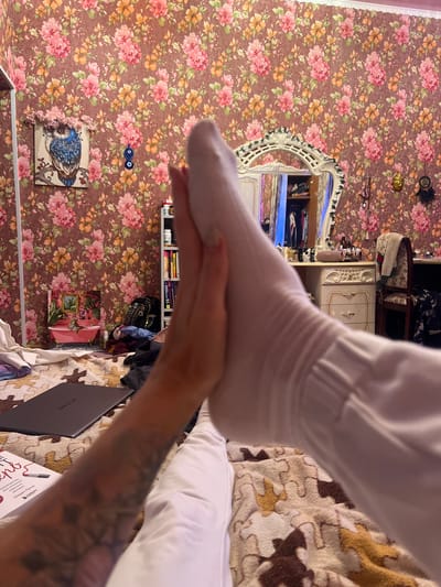 A close-up shot focuses on a person's feet and hands against a backdrop of floral wallpaper. The person is wearing a white sock on their left foot, and their hand is supporting it. The wallpaper is patterned with pink and orange flowers on a brown background. A white, ornate mirror with a carved frame is visible to the left of the person's feet. To the right, a bed with a patterned blanket and white pillows is partially visible. A laptop with the "HUAWEI" logo is on the bed. Below the mirror, a framed picture of an owl perched on a branch is hanging on the wall. Further down, another framed picture depicts a pink bathtub with a plant. In the background, shelves filled with books and various objects can be seen. The scene appears to be indoors, in a room that suggests personal living space. There is no clear indication of the time of day or weather conditions. The overall impression is casual and domestic.