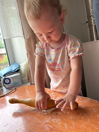 An over-the-shoulder, medium shot captures a young Caucasian toddler, with blonde hair, engaged in rolling out dough on a wooden table. The toddler, wearing a striped t-shirt with ruffled sleeves and a pink neckline, is focused on the task, their hands gripping a wooden rolling pin. Flour is scattered across the orange-toned table, indicating the ongoing activity. In the background, a window with sheer curtains reveals a glimpse of greenery and trees, suggesting an outdoor environment. To the left, on the windowsill, are several items including a box of "Отличные детки" (Excellent Kids) brand nasal aspirator replacement tips and some folded blue material. The scene appears to be set in a domestic environment, possibly a kitchen or dining area. The image is taken from a slightly elevated perspective. The location is not specified in the image itself, but if it were in Азинский, Russia, it would be a home within that city.