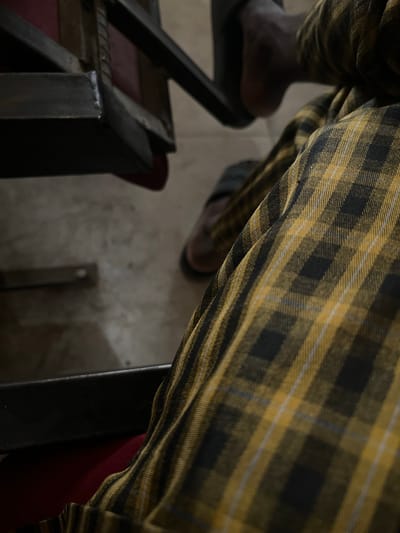 The image depicts a person wearing plaid pants in a seated position. Their dark-skinned feet are visible, one resting on the floor and the other in a black sandal atop a metal chair. A second pair of shoes is visible on the floor. The surroundings include a metal chair frame, a red seat cushion, and a grey-toned floor. The location is specified as Baganakwo, Nigeria, although there are no visual cues to confirm this.