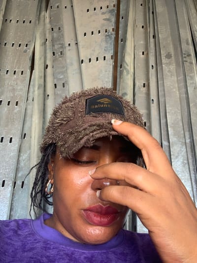 A person wearing a brown, fringed beanie with a black label is holding their hand near their face. They have dark skin, dark hair pulled into braids, and are wearing a purple top. The background consists of multiple parallel metal racks. There is no information to identify that it is Shaba, Nigeria.