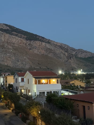 The image captures a twilight scene in Capaci, Italy, with a prominent mountain range dominating the background. The mountain slopes are rugged and sparsely vegetated, transitioning from a bare, rocky upper section to a more densely covered lower part. A clear, twilight sky with two faint points of light, possibly stars or distant aircraft, is visible above the mountain.
In the mid-ground, a cluster of houses with red-tiled roofs are nestled against the mountainside. One house, painted white, features a brightly lit upper balcony where several people are seated around a table. Other houses in the vicinity exhibit various architectural styles and colors, including yellow and white facades. Streetlights cast a warm glow on the scene, illuminating the immediate foreground and the pathways between the houses.
The foreground shows a winding road or pathway lined with thick, green bushes and trees. Some of these plants are illuminated by yellow lights, creating a dramatic contrast with the deepening twilight. Several red trash receptacles are visible along the roadside. The overall impression is a residential area on the outskirts of a town, with the imposing natural landscape serving as a dramatic backdrop.
