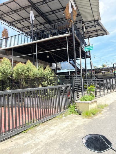 The image shows a multi-story building with an open-air second level, likely a cafe or restaurant, in the city of Langsa, Indonesia. The structure is made of metal with a corrugated metal roof. There are black metal railings on both levels, and chairs and tables are visible on the upper level. A green directional sign with white text reading "AZWA KUPI" and an arrow pointing to the left is attached to a pole near the upper level. Several small, green trees are planted behind a black metal fence in the foreground. The ground is paved with concrete and asphalt. The sky is blue with scattered white clouds. The image appears to be taken from a vehicle, as a motorcycle's rearview mirror is visible in the lower right corner.