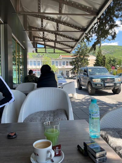 The image shows an outdoor cafe setting. A person in a black vest sits in a white wicker chair, likely at a table. Another person in a camouflage shirt sits nearby. On the table, there is a water bottle, multiple packs of cigarettes, a cup of coffee, and a glass. A vehicle, identified by the license plate "E 3001 HB", is parked on the street. The background shows buildings and a mountain. The presence of “COOP” and the Cyrillic letters indicate the location to be possibly in Eastern Europe. Given the context cues from the Cyrillic characters on the water bottle (Velingrad) and cigarettes (Sobranie), this location is in the city of Sopot, Bulgaria.