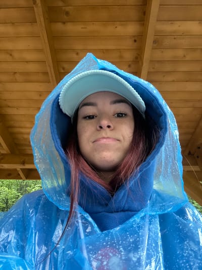 A young woman stands under a wooden structure, likely a gazebo or pavilion, in Pejë, Kosovo. She wears a blue hooded sweatshirt, a light blue visor, and a clear blue plastic poncho, presumably for rain protection. The background reveals trees, suggesting an outdoor setting, potentially a park or green space. The woman's gaze is directed towards the camera with a slight smirk.