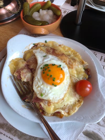 A medium-shot photograph depicts a dish of Reblochon cheese tartiflette topped with a fried egg, a tomato, and chives, placed on a white plate that rests on a table with a newspaper placemat. A fork partially obscures the food. A small orange bowl containing pickles and small white onions sits beside the plate. A portion of a black object and a copper-colored teapot are visible to the side of the bowl. The words "orge de la Halle" are visible on the newspaper placemat. The scene is set in Epagny, Switzerland.