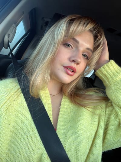 A fair-skinned female, appearing to be a young adult, is depicted seated within the passenger compartment of a vehicle. She has blonde hair styled with bangs and is looking directly at the camera. Her left hand is positioned with fingers near the back of her head, resting lightly on her hair. She wears a lime green knitted cardigan over a visible pink undergarment. A dark-colored seatbelt crosses diagonally from her left shoulder to her right hip. A thin, light-colored necklace is visible around her neck. Portions of the vehicle's dark interior and a light-colored overhead lining are visible in the background. The specific location in the city of Азьмушкино, Russia, cannot be identified from the visual content of this image.