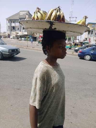 A young person stands in profile on a paved road in Mumbwa, Zambia, under a clear, bright sky. The individual, with dark skin and short, dark dreadlocks or braids, wears a light-colored, textured, short-sleeved knit shirt. They balance a large, round, light-colored metal tray on their head, which contains several bunches of ripe bananas, some with dark spots. The person faces right, looking forward with a neutral expression. In the left background, a light-colored sedan is partially visible on the road, with a multi-story building and other structures further behind. The right background reveals a street scene with additional vehicles, including a blue sedan and a three-wheeled auto-rickshaw, along with various buildings and a tall metallic communication tower. Several indistinct figures are visible in the distant background.