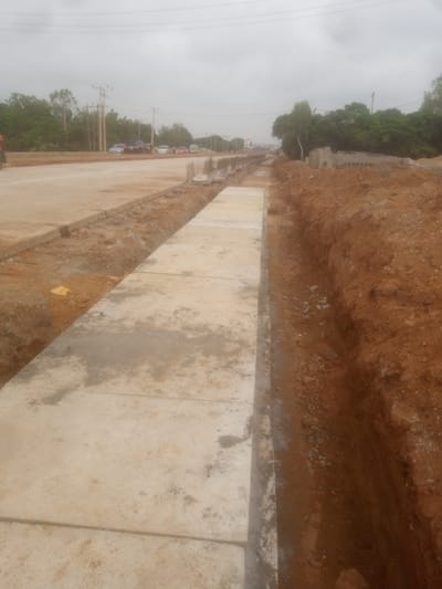 This image depicts a construction site in Kakuri Gwari, Nigeria, with a focus on ongoing roadwork. A newly constructed concrete sidewalk runs along the right side of the frame, bordered by a deep trench filled with loose soil and rocks. The ground adjacent to the sidewalk appears to be leveled and prepared for further construction. In the background, a road is visible with several vehicles, including cars and what appears to be a truck, suggesting active traffic. Utility poles and power lines are present, indicating an urban or semi-urban setting. Trees and vegetation line the horizon, partially obscured by an overcast sky. The scene conveys a sense of development and infrastructure improvement in progress.
