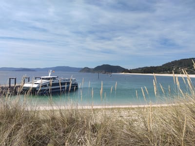 The image depicts a scenic coastal view in the city of Vigo, Spain, likely from a beach. In the foreground, dry grass and reeds line the sandy shore. The calm, turquoise water of the bay extends towards the horizon, where rolling hills covered in vegetation form the distant landscape. A modern ferry boat, emblazoned with the text "mar de ons", is docked at a pier on the left side of the frame. Several passengers can be seen on the boat and on the pier. In the middle ground, other boats are anchored in the bay, and buoys dot the water. The sky is a vibrant blue, with scattered white clouds. The overall impression is one of a peaceful, sunny day at a popular coastal destination.