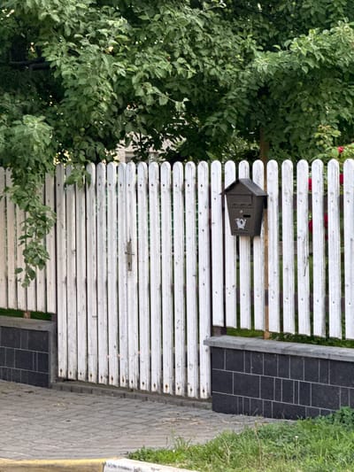 The image depicts a white picket fence with a dark mailbox attached to it. The fence is weathered, with some rust visible on the pickets. A dark, brick-like structure forms the base of the fence on the right side of the image, topped with a grey stone cap. Behind the fence, lush green trees are visible, and a hint of red flowers can be seen on the right. The foreground shows a paved walkway with a yellow curb on the left and some green grass. The scene is likely a residential area in Irpin, Ukraine, given the context. No people are present in the image, and no specific actions or interactions are taking place. The overall environment suggests an outdoor, possibly suburban setting.