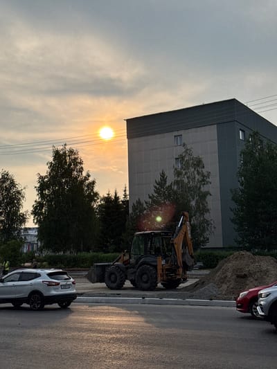 The image depicts an outdoor scene in Surgut, Russia, likely during the late afternoon or early evening, given the low sun casting a warm glow and creating hazy conditions. The primary focus is a construction site with a yellow CASE backhoe loader in the foreground. The loader is parked on a wet asphalt surface, suggesting recent rain or damp conditions.
In the background, a large, modern building with a grey facade dominates the left side of the frame. Trees with dense green foliage surround the building and the construction area. A pile of dirt and gravel is visible next to the building, further indicating ongoing construction work.
Several vehicles are present: a red car and a silver car are parked closely together on the right side of the image, partially obscuring a mound of dirt. A white SUV with a license plate starting with "P808" is parked on the asphalt in the lower right quadrant of the image.
The sky is overcast with thin clouds, through which the sun is visible as a bright, diffused orb, creating a lens flare effect. The overall atmosphere is somewhat muted due to the lighting and weather conditions. There are no clearly discernible people in the image, though it's possible some are obscured by the machinery or vehicles. The scene conveys a sense of routine work in progress, with the natural light adding a touch of warmth to the industrial setting.