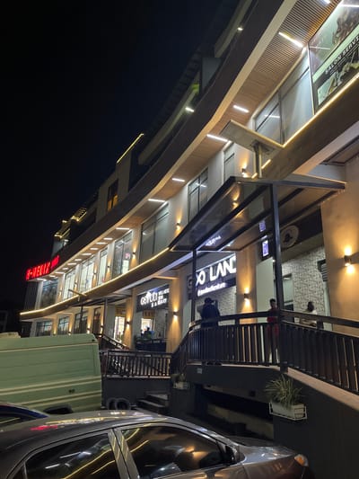 This is a nighttime photograph of a modern building with multiple levels, illuminated by warm, indirect lighting. The building appears to be a commercial establishment, likely housing various businesses. On one of the upper levels, a sign reads "SALON SERVI," suggesting a salon or spa. Another prominent sign further down the building displays "H-MEDIX," possibly indicating a medical or pharmaceutical business. Above this, a sign reads "GOOGLANE WELLNESS SKIN SA BEAUTY," advertising beauty and wellness services. The building's architecture is sleek, with glass facades and contemporary detailing.
In the foreground, a silver car is parked, its headlights and reflections adding to the nighttime ambiance. A portion of a light green shipping container is also visible. The overall scene depicts an urban commercial area at night, with businesses illuminated and ready for operation. The presence of multiple signs suggests a vibrant retail or service hub. The lighting creates a sophisticated and inviting atmosphere, despite the darkness of the night. No specific activities or emotions are clearly depicted, but the well-lit storefronts imply a sense of business activity. The location context of Guzape, Nigeria, places this scene within an African urban setting.