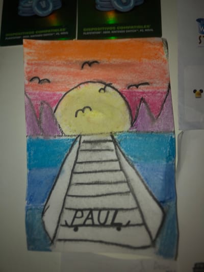 The image shows a drawing on paper, likely made with crayons or colored pencils. The drawing depicts a stylized sun with a smiley face, situated on the left side of the frame. The sun is colored yellow and has simple black lines for eyes and a mouth. In front of the sun, there are two small, black, curved shapes that could represent birds.

The background of the drawing features a vibrant gradient of colors, transitioning from orange and pink on the left to a bright blue on the right. This suggests a sunset or sunrise scene. On the right side of the drawing, a structure that resembles a staircase or a ramp is drawn in black and white. The word "PAUL" is written vertically along this structure.

The drawing is placed on a surface that appears to be a table or shelf. To the left of the drawing, there are two dark green boxes with holographic text that reads "DISPOSITIVOS COMPATIBLES PLAYSTATION, XBOX, NINTENDO SWITCH™, PC, MOVIL" in multiple languages, indicating they are related to gaming or electronic devices. Above the drawing, a piece of paper with a blue-tinted background and some text is visible.

The overall impression of the drawing is childlike and cheerful, with the bright colors and simple representation of the sun conveying a positive emotion. There are no specific cues about the time of day, weather, or precise location within l'Hospitalet de Llobregat, other than the general context of the gaming device boxes. The drawing appears to be a personal creation, possibly a gift or a doodle.