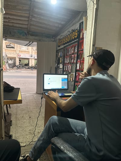 A man wearing a light blue polo shirt and blue jeans sits at a desk in what appears to be a small shop, looking at a laptop. He wears a black baseball cap. The laptop screen displays a "Your connection was interrupted" message. The shop has a partially exposed wooden ceiling and shelves displaying various phone accessories. Through an open doorway, a street scene is visible with buildings, a pile of tires, and a person walking in the distance. The overall environment suggests a modest business location in a developing urban area. The text on the screen is English. The text on the signage in the shop is partially visible and appears to be in English and possibly Arabic. The location is described as Gaza, Palestinian Territories.