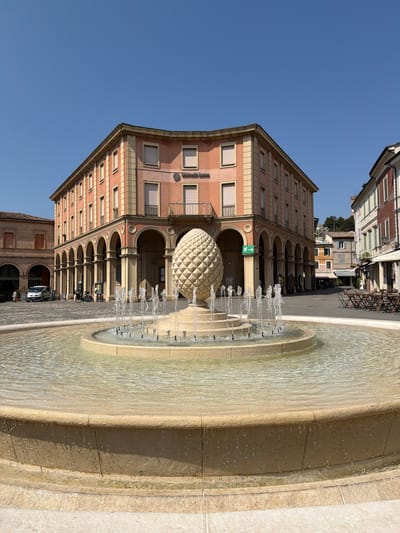 This is a full-shot, eye-level, outdoor photograph taken in the city of Santarcangelo di Romagna, Italy, on a clear, sunny day. In the foreground, a multi-tiered, round fountain with a stone basin is active, with water spraying upwards. A large, textured, light-colored sculpture resembling a pinecone or artichoke stands in the center of the fountain. Behind the fountain is a cobblestone plaza. Dominating the mid-ground is a light salmon-colored building with a curved facade, featuring a ground floor arcade with arched walkways and many windows on the upper floors. The building has an "UniCredit Banca" sign visible. To the left of this building is a brick structure with arched openings, and a silver car is parked in front of it. To the right of the salmon-colored building, other buildings line the plaza, one with red brick and arched windows, and another with a white awning and outdoor seating. The sky is a clear, vibrant blue. There are a few people visible in the scene, but they are small and not the primary focus.