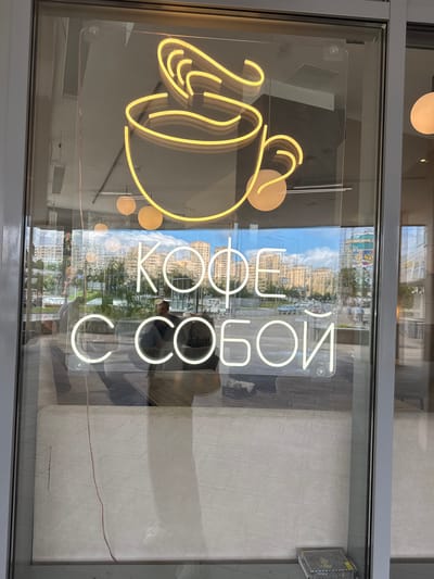 The image shows a neon sign in Russian displayed on a glass storefront, likely in the city of Zelenograd, Russia. The neon sign features a graphic of a steaming coffee cup and the text "КОФЕ С СОБОЙ" which translates to "Coffee to go". The sign is illuminated in yellow and white neon. Through the glass, a street scene is visible with apartment buildings, cars, and pedestrians. Inside the establishment, the reflection of the street and sky can be seen on the glass. A person is partially visible inside, and there is furniture and some equipment on the floor.