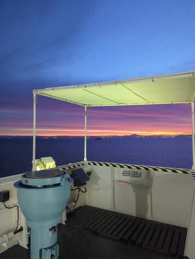 The image captures a view from the deck of a vessel at sea, possibly a ferry or cargo ship, looking towards the coastline at twilight. The sky displays a vibrant gradient of blues, purples, and oranges, indicative of sunset or sunrise. In the foreground, a large, light blue marine compass housing is prominent, with associated electrical boxes and wiring. A striped yellow and black railing runs along the edge of the deck, with height markings (16.30 M / 53.48 FT and 183.66 M / 602.56 FT) visible on the white structure behind it. A dark, slatted mat covers part of the deck. Beyond the railing, a calm sea stretches to the horizon, where a distant city skyline is illuminated by lights. The coastline features rolling hills or mountains. The city lights and the overall landscape suggest an urban or industrial area on the coast of Yokkaichi, Japan, known for its port and industrial complexes. The presence of the sea, the navigational equipment, and the distant city lights establish the context of maritime activity near an urban center. There are no people visible in the image.