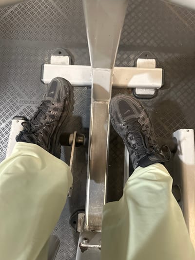 The image shows a person’s feet resting on the footrests of a rowing machine. The person is wearing grey running shoes and light-colored pants. The rowing machine is made of white metal and has a diamond-patterned floor beneath it. The location in the city of Tunbridge Wells, United Kingdom, cannot be determined from the visual information.