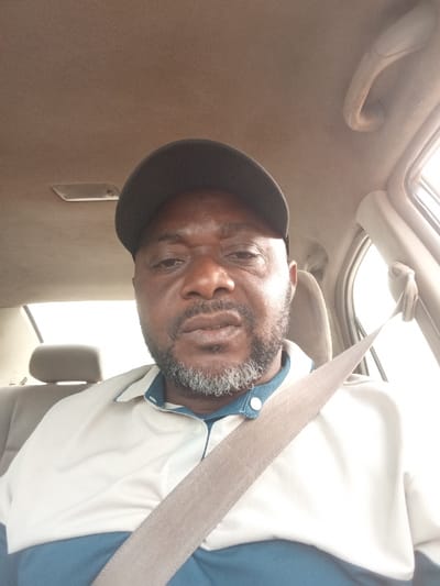 A male subject is depicted inside a vehicle. The scene is identified as being within the city of Peyer, Nigeria. The male has dark skin, a dark baseball cap covering his head, and a graying beard and mustache. He wears a polo shirt, cream-colored on the upper torso with a collar and dark teal below, fastened with white buttons at the collar placket. A light brown seatbelt crosses his chest diagonally from the upper right to the lower left. The vehicle's interior features a light-colored headliner, an overhead light fixture, and a grab handle on the right side. The subject's gaze is directed towards the viewer/camera.