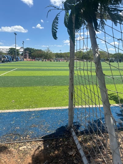 This image captures a vibrant outdoor sports field on a clear, sunny day, likely in Mérida, Mexico. The perspective is from directly behind a soccer goal, looking out across the field through the white mesh netting.

The foreground is dominated by the goal's white metal posts and the netting, which creates a patterned overlay on the scene. To the left, a dark, leafy tree branch partially frames the view. To the right and below the goal, the ground consists of dry dirt and scattered leaves, contrasting with the manicured field.

The main subject is a large artificial turf field, characterized by alternating stripes of light and dark green, indicating a playing surface. A clear white line marks the boundary of the green turf, adjacent to a bright blue perimeter or track that encircles the field.

In the mid-ground and background, several people are visible on the field, appearing to be engaged in some form of athletic activity or gathering, though their specific actions are not discernible due to the distance. Beyond the field, a line of lush green trees stretches across the horizon. Overhead, the sky is a brilliant blue with scattered, fluffy white clouds. Several tall light poles with fixtures are also visible against the sky, suggesting the facility can be used at night. No distinct text is readable on any signs or banners in the distance.

The overall impression is one of a well-maintained sporting venue under bright, clear weather conditions.