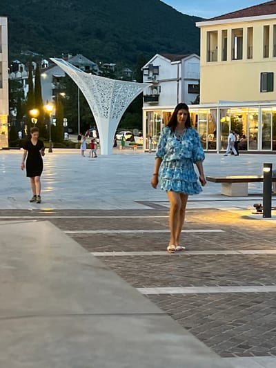 This media file captures an outdoor scene in Kumbor, Montenegro, likely during dusk as indicated by the ambient lighting and the transition from daylight to evening illumination from street lamps and building interiors.
The primary subjects are two individuals. In the foreground, a woman wearing a short, blue, patterned dress and white sandals stands on a paved walkway. She is facing the camera with a neutral expression. Further in the background, another person, dressed in a black dress and sneakers, is also standing on the walkway, facing away from the camera.
The setting appears to be a modern waterfront area. Behind the woman in the blue dress, a light-colored building with multiple windows, possibly a hotel or restaurant, is visible. The windows reflect the interior lights and show glimpses of other patrons. In the middle ground, a striking white, abstract structure with a patterned canopy is a prominent feature, likely an architectural element for shade or decoration in a public space. Beyond this, a hillside covered with lush greenery and dotted with houses slopes down towards the water, which is not clearly visible but implied by the setting. The paving stones on the walkway are arranged in a diagonal pattern, with some areas of lighter stone interspersed.
The overall mood is relaxed and casual, suggesting a pleasant evening outing. The time of day suggests the end of a warm day, with the evening lights beginning to illuminate the surroundings. No visible text is present in the image.