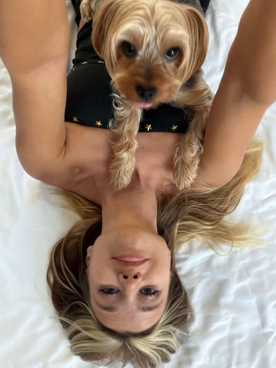The media file features a close-up, overhead shot of a woman lying on a white surface, possibly a bed, with a small, light brown dog nestled beside her. The woman has blonde hair with lighter streaks and is looking towards the camera. She is wearing a black garment adorned with gold stars. The dog, a Yorkshire Terrier, is positioned in the foreground, with its face and paws visible. It appears to be looking up at the camera, with its tongue slightly out. The scene is lit by natural light, suggesting daytime, and the overall mood appears relaxed and intimate. The white surface the subjects are on creates a clean, uncluttered background, emphasizing the subjects. There is no visible text in the image. The location is identified as Thep Krasatti, Thailand.