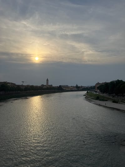 This image depicts a wide river flowing through the city of Verona, Italy, during what appears to be either sunrise or sunset. The sun is visible as a bright orb low in the sky, partially obscured by clouds, casting a golden reflection on the water's surface.

The left bank of the river shows a treeline with buildings beyond, including a distinctive tower with a clock face. The right bank features a paved walkway and a rocky shoreline where two individuals are visible walking. The river itself is calm, with small ripples indicating a gentle current. A bird is captured in flight above the water. The overall atmosphere is serene and somewhat muted due to the cloudy sky. The perspective suggests the photo was taken from a bridge overlooking the river and the cityscape.