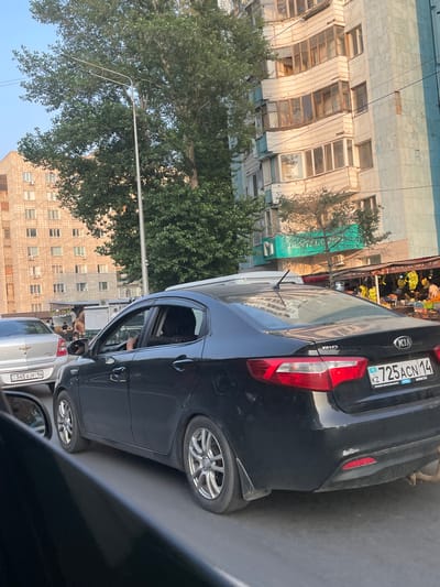 This is a photograph taken from inside a car, looking out at a street scene. The primary subjects are several cars, including a black Kia Rio in the foreground, and the facades of buildings. The scene appears to be an urban environment, likely a street or parking area.
The black Kia Rio is parked and its rear is visible, along with the license plate which reads "KZ 725ACN 14". There's also a smaller plate with "aster.kz" and Russian text. The car's taillight and part of its wheel are visible. In front of the Kia, a white Chevrolet is partially visible with its license plate reading "345 AJF 14". Another car's side is visible on the right edge of the frame.
The background features buildings with multiple windows and air conditioning units. Some of these buildings appear to be older, with weathered facades. A tree with dense green foliage is visible between the cars and the buildings, suggesting a warm season. Above the tree, power lines are strung across the sky. A street lamp is also present.
In the distance, there's a market stall or shop with fruits and vegetables displayed. Text on the buildings and market stall is in Russian and possibly Kazakh. Examples of visible text include "KIA", "RIO", "ЖЕТКІЗУ ДОСТАВКА", and phone numbers.
The lighting suggests it is daytime, possibly late afternoon given the angle of the light. The overall impression is of a busy street in a city, with parked vehicles and everyday urban life. There are no prominent displays of emotion. The location cues like the license plates and text point to Kazakhstan, specifically Pavlodar.