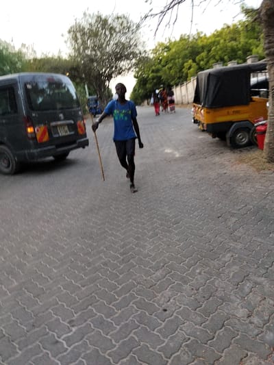 The scene depicts a paved road in Gombato Bongwe ward, Kenya, under diffused natural light, consistent with late afternoon or early evening. The road surface is constructed from grey interlocking concrete pavers laid in a herringbone pattern.

In the central foreground, a dark-skinned individual, wearing a blue short-sleeved t-shirt and dark trousers or shorts, is observed moving towards the viewer's left. This individual holds a long, slender, light-brown wooden stick in their right hand, angled downwards.

To the left, a dark-grey minivan, possibly a Toyota Hiace or similar, is partially visible from the rear, displaying illuminated red taillights and orange turn indicators. A Kenyan registration plate, partially obscured, is affixed to its rear.

To the right, a yellow motorized rickshaw (tuk-tuk) equipped with a black canvas canopy is positioned, displaying "3 PASS" on its side. A section of a red vehicle, possibly a motorcycle or scooter, is partially visible behind the tuk-tuk. Further down the road, on the right, a group of approximately three to five individuals are walking away from the viewer.

The environment is characterized by lush green foliage from trees lining both sides of the road. A light-colored wall runs along the right side of the road, behind the tuk-tuk and the pedestrians. The sky overhead is pale and hazy. A tree trunk is visible in the extreme right foreground.