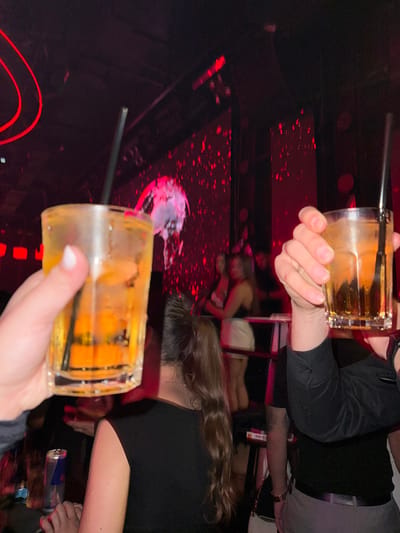 The image depicts an indoor scene within a darkly lit establishment, identified as a nightclub or bar in Tirana, Albania. The primary focus is on two individuals' hands in the foreground, each holding a clear, faceted glass containing an amber-colored liquid with ice cubes and a black straw. The glass on the left is held by a hand with manicured fingernails painted white. The glass on the right is held by a hand belonging to an individual wearing a black long-sleeved shirt.
In the mid-ground, below the left glass, the back of a female's head and shoulders are visible. She has long, dark brown hair styled in a high ponytail and is wearing a black sleeveless top. A thin silver necklace is discernable on her neck. To her left, a silver Red Bull can rests on a surface. Below the right glass, part of a male's torso is visible, clad in a black short-sleeved shirt over a darker garment, paired with light-colored trousers and a black belt. A watch is present on his left wrist.
The background is characterized by dim red illumination. A large screen displays abstract red patterns, including what appears to be a circular or spherical object with emanating splashes or particles. Several blurry figures, predominantly female, are scattered in the background, some appearing to stand near a raised platform or bar. Red linear lighting elements are visible on the ceiling. The overall environment suggests a social gathering at night.