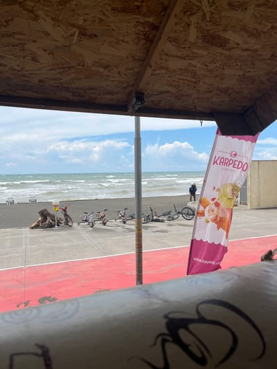 This image captures a scene near the Black Sea coast in Ureki, Georgia. In the foreground, a wooden structure with visible wood chips forms a canopy overhead, partially obscuring the view of the environment beyond. A banner with the text "KARPEDO DONDURMA" and a website address "www.karpedo.com" is prominently displayed on the right side of the frame, featuring images of ice cream.

The middle ground shows a paved area with a vibrant red surface and white lines, suggesting a recreational court. Several electric scooters or similar recreational vehicles are parked on the paved area. To the left, a large piece of driftwood rests on the pavement near a gumball machine. Beyond the paved area lies a dark sand beach, where two empty lounge chairs are set up facing the sea. The Black Sea stretches out with moderate waves under a sky filled with white, puffy clouds. A single individual is visible on the beach, standing near the water and appearing to be holding a stick or pole.

In the background, a light-colored building with vertical supports is partially visible on the right. The overall environment suggests a seaside recreational area, likely a promenade or park adjacent to the beach. The image is taken from a slightly elevated perspective, looking down towards the beach and the sea.