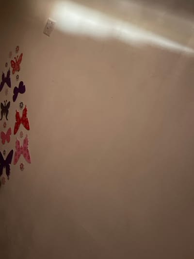 This image displays an interior wall in Tamaha, Nigeria. The wall is light-toned, possibly beige or off-white, exhibiting minor surface variations. A distinct horizontal band of bright light extends across the upper right section, indicating an overhead light source. In the upper-left quadrant, a white electrical wall plate is affixed. The left side of the wall is decorated with a vertical arrangement of adhesive butterfly cutouts in various sizes and colors, including red, purple, dark brown, and pink. Smaller, patterned floral cutouts are interspersed among the butterflies. No human subjects or active events are depicted.
