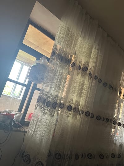 The image depicts an interior scene with a window covered by a sheer, patterned curtain. The curtain is white with a decorative, dark blue floral motif and intricate geometric patterns. Sunlight filters through the curtain, creating shadows and highlighting the fabric's texture.

Through the window, a glimpse of the outdoors is visible. The window frame appears to be made of dark material, possibly metal or wood. Outside, there are trees with green foliage, suggesting a natural environment.

On the windowsill, several objects are present. A white plastic bag, seemingly filled with various items, hangs from a support structure near the window. Electrical cords and a red electronic device are also visible on the windowsill, suggesting some form of equipment or technology is set up there. The wall around the window appears to be constructed of concrete blocks, some of which are covered with a white material.

The overall impression is of a domestic or working space, possibly a room in a building under construction or renovation, given the visible concrete and the unfinished appearance of the window area. The lighting suggests it is daytime. The location is identified as Mıxlıqovaq, Azerbaijan.